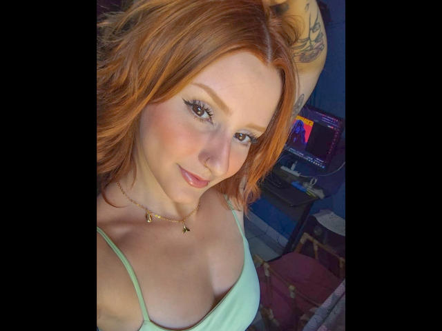 RuivaK with red hair, green top, small tits, tattoo on arm, subtle smile, light makeup, necklace, and cute face, looking at camera.