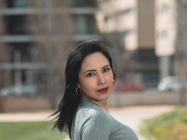 ValeriaQ with dark hair, red lipstick, gray ribbed shirt, small hoop earrings, looking over shoulder, slight smile, blurred urban background. Boobs not visible, focus on her face.