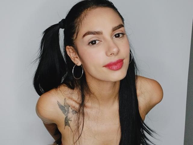 AnaisHilton with black pigtails, hoop earrings, red lipstick, and a bird tattoo on her right shoulder, smiling at the camera. Her boobs are partially visible.