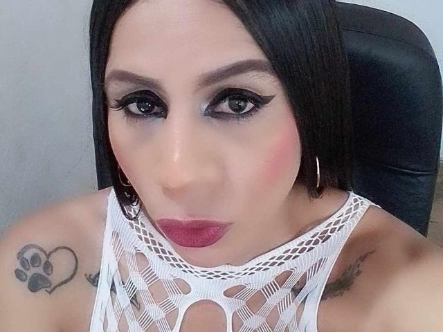 TrayXibi with dark hair, heavy eyeliner, pink blush, red lipstick, white fishnet top, small tits, hoop earrings, paw tattoo on left shoulder, pouty expression, black chair.