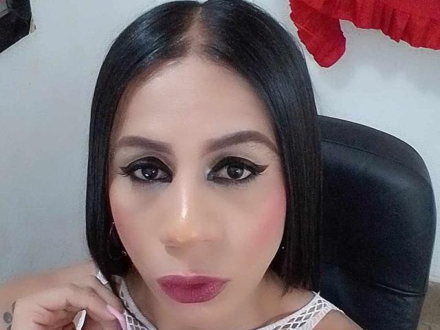 TrayXibi has straight black hair, thick eyeliner, dark eyebrows, pink lipstick, and a white lace top. She's looking at the camera with a serious expression.