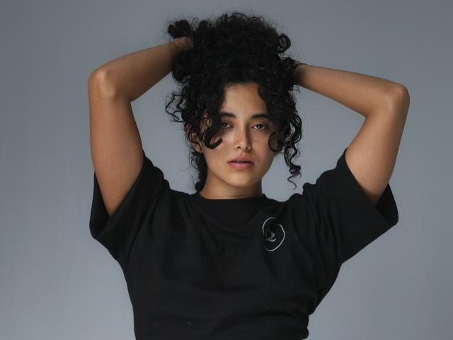 Vivienne Laurent mit lockigem, schwarzem Haar, mit erhobenen Armen, trägt ein schwarzes Hemd und blickt direkt in die Kamera, grauer Hintergrund. Ihre Brüste sind klein, und sie trägt dunkles Make-up unter den Augen..