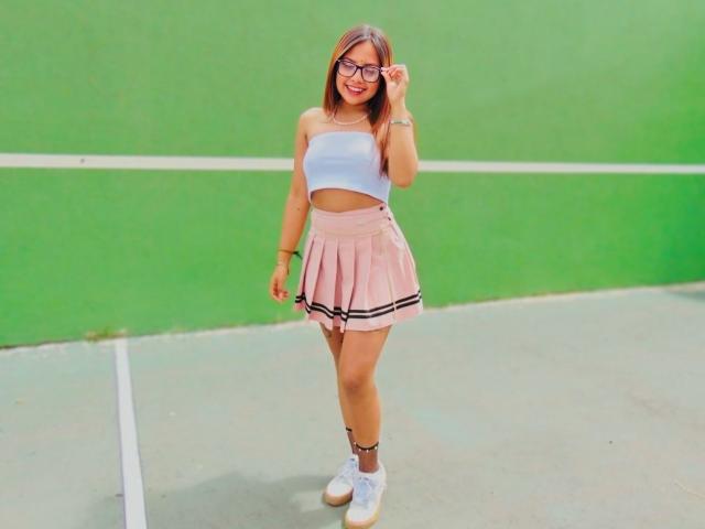 NinaDeville has brown hair, glasses, a blue crop top, pink pleated skirt with black stripes, and white sneakers, standing on a green tennis court.