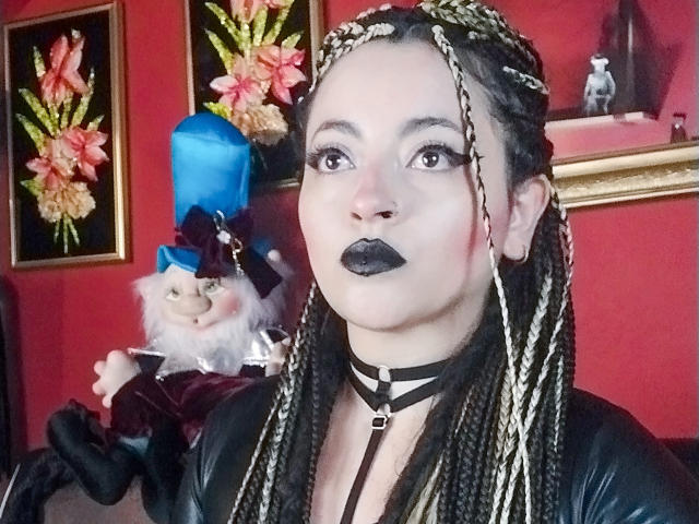 LunaTikka with braided black and blonde hair, black choker, black lipstick, and leather top, holding a doll with a blue hat, in a red room with floral pictures.