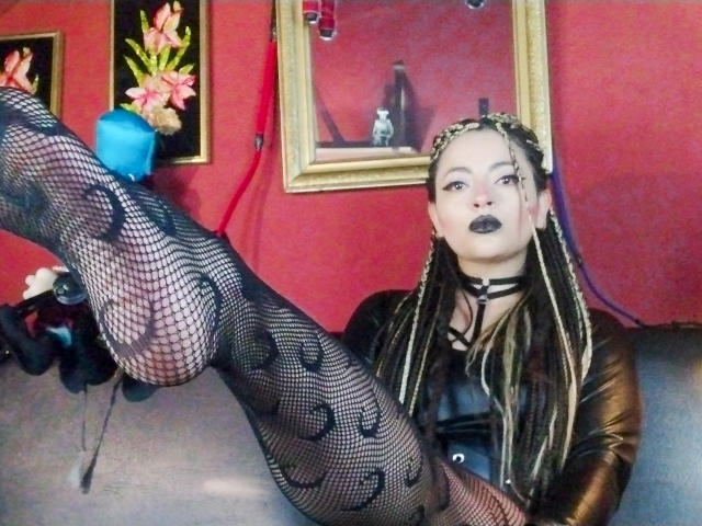 LunaTikka with black fishnet stockings, patterned, and raised leg showing her foot, wearing a black leather top, dark lipstick, and braided hair, in a red-walled room.