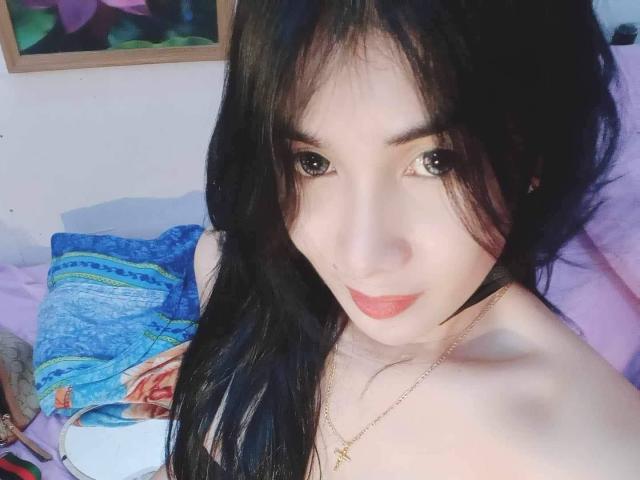 Iyutlame with long black hair, pale skin, and red lipstick, looking at the camera with big eyes, wearing a gold necklace, and no top, showing her bare shoulders.