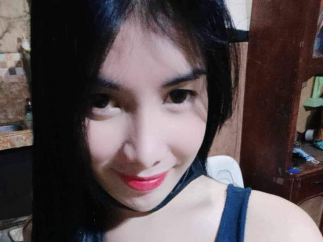 Iyutlame with dark hair, red lipstick, and black tank top, smiling at the camera, showing her smooth, light-skinned face and small, round nose. Her tits are not visible.