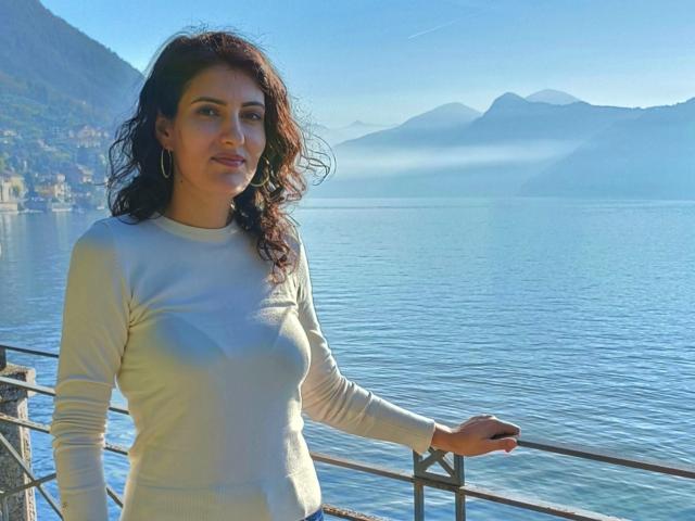 AddictedToUTS with wavy brown hair, wearing a white long-sleeve shirt, standing by a railing, looking at the camera, with blue water and misty mountains in the background.
