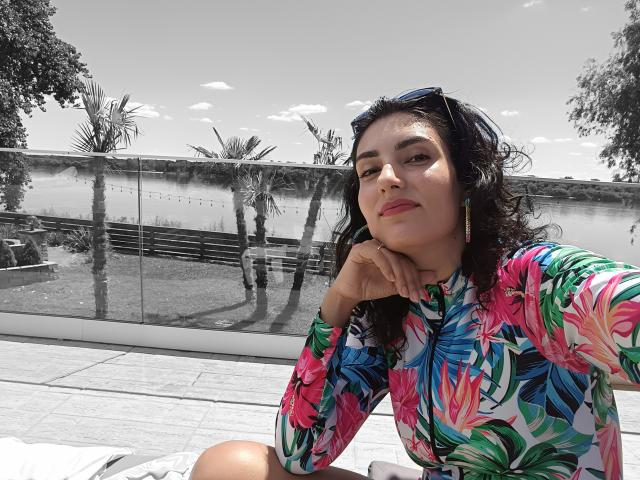 Viciado em UTS, com camisa floral colorida, cabelo ondulado escuro, batom rosa, óculos de sol na cabeça, mão no queixo, sentado ao ar livre, fundo preto e branco, luz do dia forte, expressão confiante..