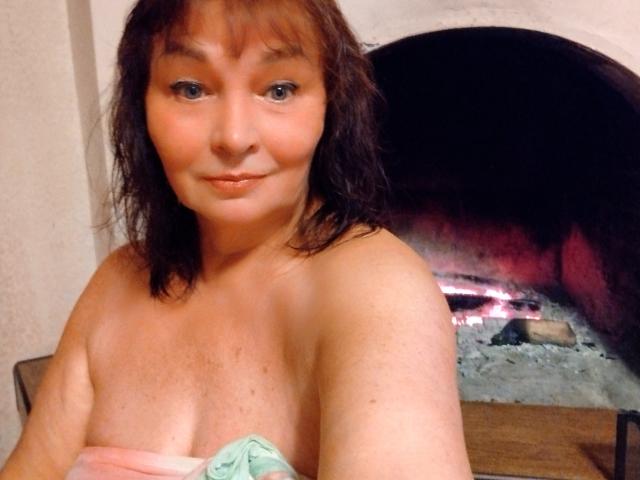 MilaMils with shoulder-length brown hair, blue eyes, and a subtle smile, topless, covering her small, perky tits with a green cloth, in front of a lit fireplace.