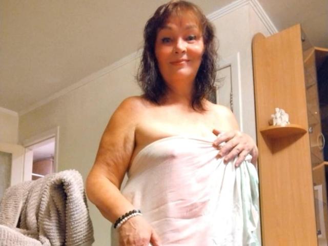 MilaMils with wavy brown hair, medium skin, and a towel covering her tits, holding a blue towel, smiling in a room with a wooden door and shelf.