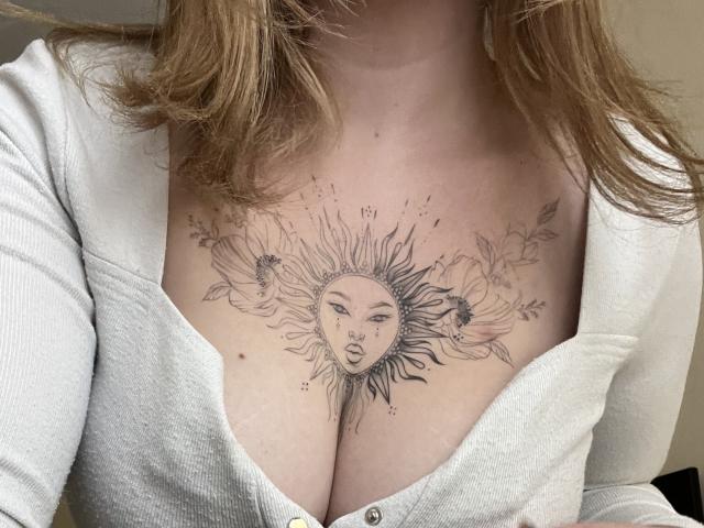 DiLaria's chest tattoo features a sun face with a woman's eyes and open mouth, surrounded by flowers and leaves, just above her tits. She's wearing a white cardigan.