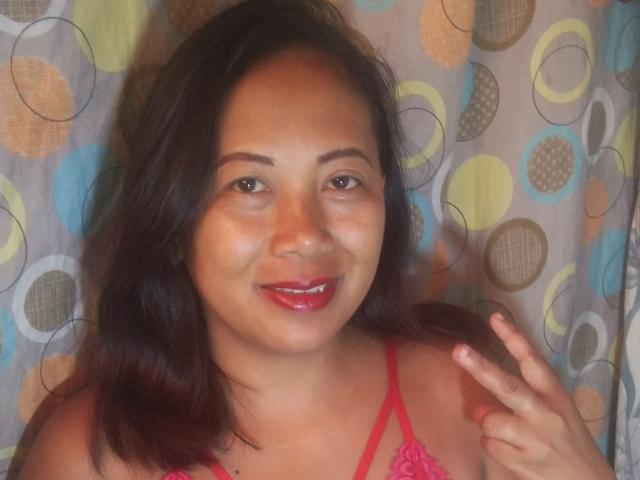 SarahFontaineX with dark brown hair, red lipstick, and pink lace top, smiling at the camera, making a peace sign with her right hand. She's got cute tits.
