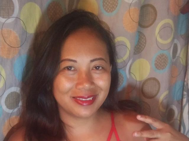 SarahFontaineX with dark brown hair, red lipstick, and a smile, wearing a red top, against a colorful patterned curtain. Her hand is raised near her face.