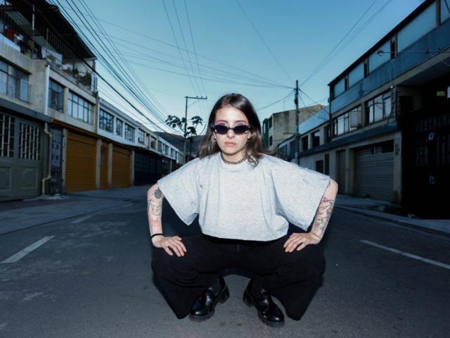 AmySpider squatting on a street, wearing a gray shirt, black pants, black boots, and sunglasses, with tattoos on her arms and hands, looking cool and confident.
