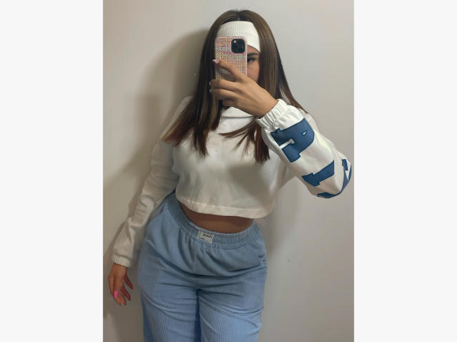 LunaMiranda with medium tits, wearing a white crop top and blue pants, taking a mirror selfie, white headband, long brown hair, white wall background, phone in hand.