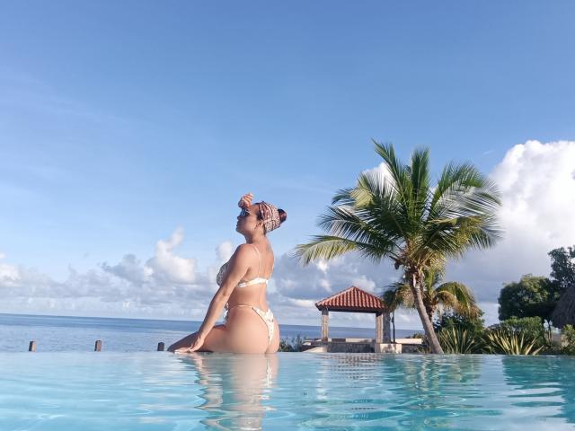 GalaMalkova in a white bikini, sitting in a pool with blue water, facing sideways, looking up, her ass and tits visible, a palm tree and red-roofed shack in background.