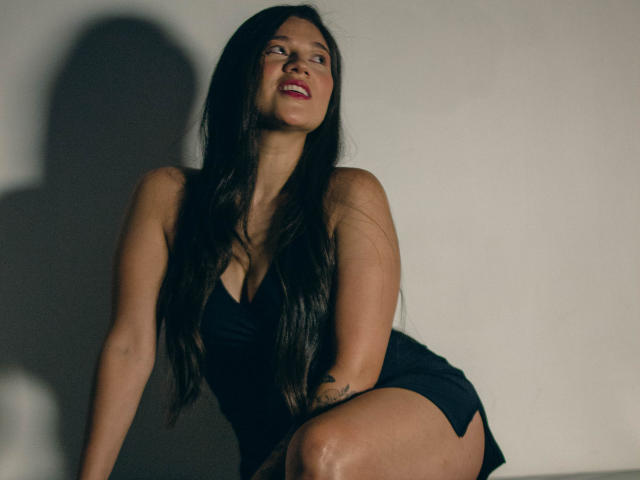 LindaJimenex, com cabelos pretos longos, sorrindo, vestindo um vestido preto, mostrando o bumbum, em pé contra uma parede sombria, olhando para cima, batom vermelho, tatuagem no braço direito..