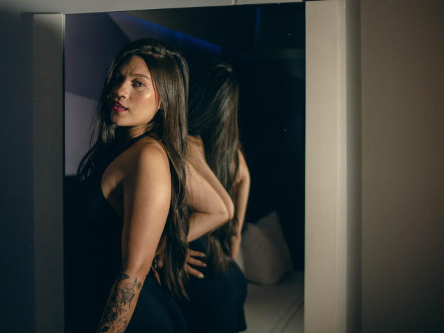 LindaJimenex met lang bruin haar, een zwarte jurk, een tatoeage op haar rechterarm, die in de spiegel kijkt. De reflectie toont haar rug en billen. Donkere, sfeervolle belichting. Zacht, sensueel en zelfverzekerd..