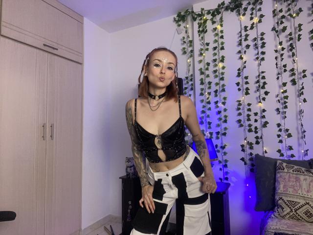 SarahRousen with tattoos, brown hair, wearing a black cutout top, white and black pants, choker, and nose ring, pouting in a room with leaf garlands and blue lights.