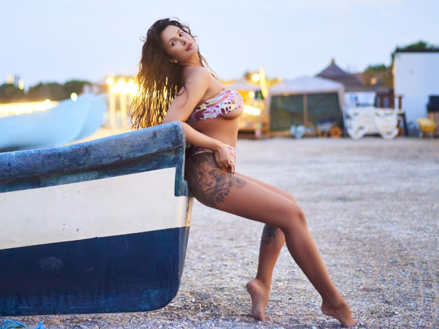 JaneDivine with long brown hair, wearing a floral bikini, leaning on a blue-and-white boat, showing a large tattoo on her right thigh, standing barefoot on a gravel beach.