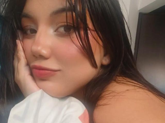 SquirtinGamy with wet black hair, resting cheek on hand, soft pink lips, slight smile, brown eyes, fair skin, subtle blush, white shirt with pink accents, relaxed expression.