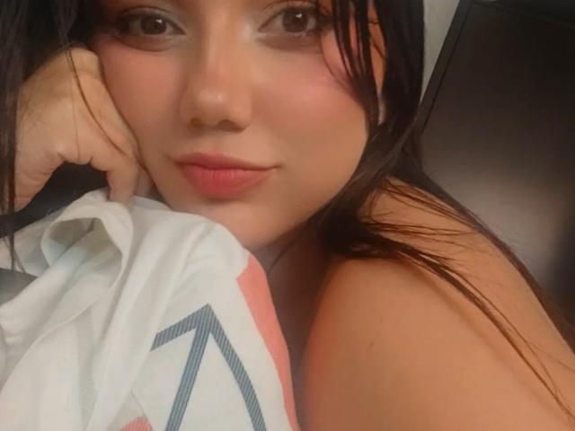 SquirtinGamy with long black hair, slightly flushed cheeks, and pink lips, lying on a bed with a white and pink patterned blanket, covering her tits. Her hand rests on her face.