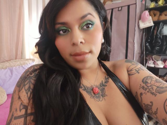 Chili con sombra de ojos verde y tatuajes, usando un cabestro de cuero negro, recibiendo un footjob, mirando a la cámara.