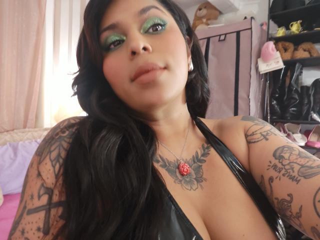 Chili, com longos cabelos pretos, sombra verde, e tatuagens, está vestindo um top de couro preto, mostrando grandes mamas e olhando olhando para a câmera.