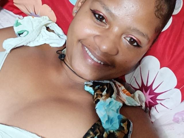 MeriAndMark with dark brown skin, smiling, wearing a floral dress with visible cleavage, black necklace, and subtle eyeliner, lying on a red and white floral pillow.