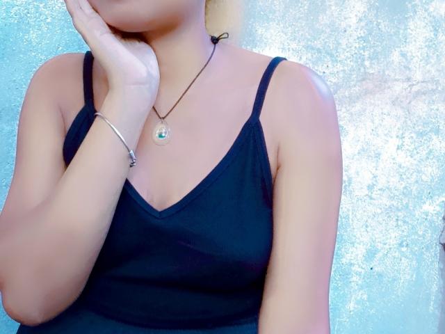 MeriAndMark has tan skin, wears a black tank top, a silver bracelet, and a necklace with a green gem, touching her cheek with one hand.