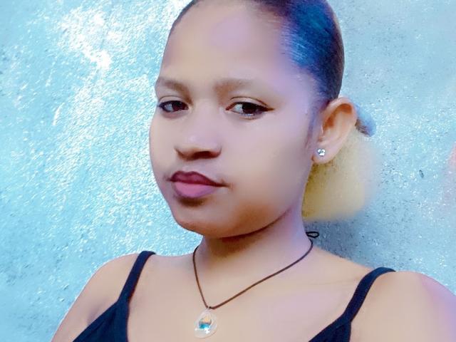 MeriAndMark has medium brown skin, black hair in a ponytail, small earrings, dark lipstick, and a black top with a necklace. Her tits are not visible.