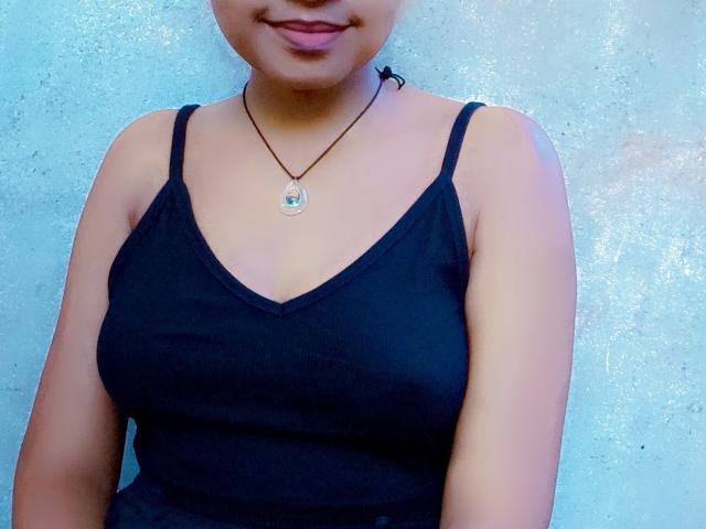 MeriAndMark has big tits in a black tank top, a necklace with a green pendant, and a subtle smile, standing against a textured blue wall.