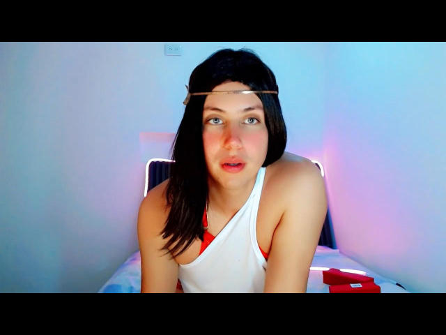 AdriudI with black hair, wearing a white tank top, has a gold headband, and is looking to the side with red underwear visible, sitting on a bed.