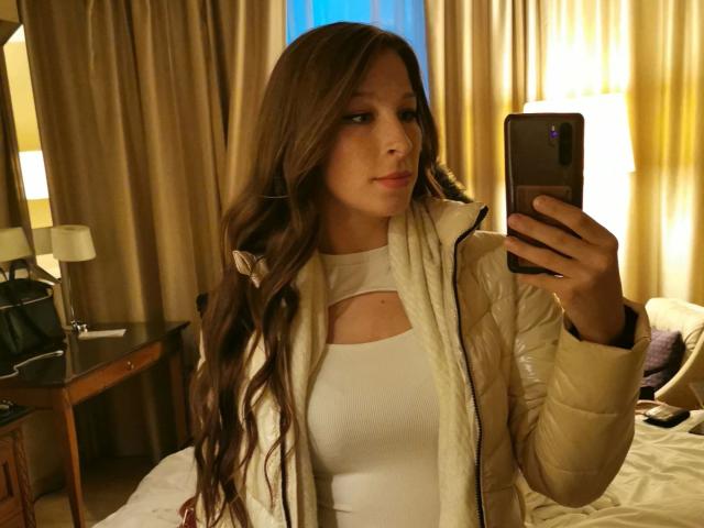 Livie-hot with long brown hair, white top, and puffy jacket, holding a phone, taking a mirror selfie, showing her tits and a small cutout in her top.