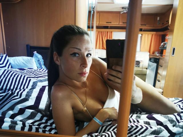 Livie-hot with brown hair, wearing a necklace, lying on a striped bed, taking a mirror selfie, showing her small tits and a white towel covering her pussy.