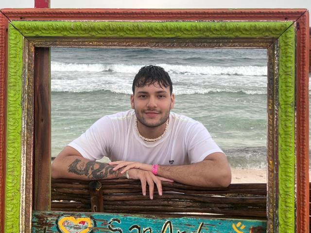HenryEdward with short dark hair, white t-shirt, tattoo on right arm, pink bracelet, and bead necklace, leaning on wooden frame, green and red ornate border, beach waves behind.