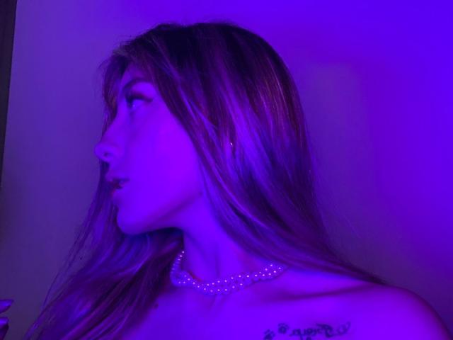 LivelyVanderWood with long, straight hair, purple-lit profile, side-eye, necklace, and tattooed shoulder, looking off to the side. Her tits are slightly visible.