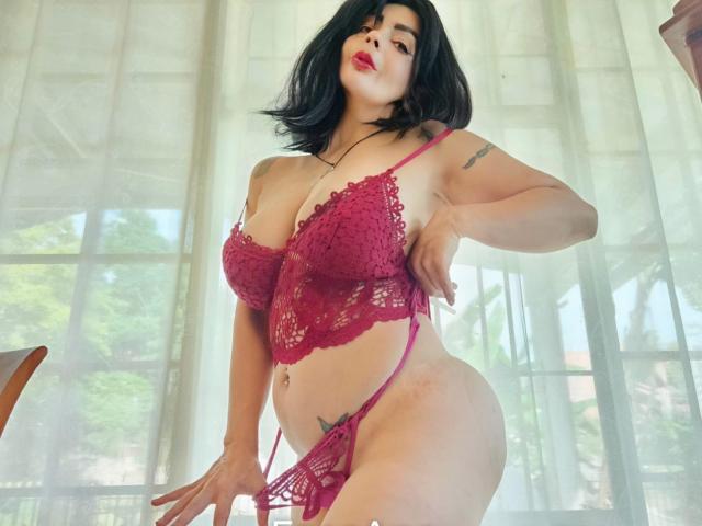 BrandyMostHottest in red lace lingerie, showing her big tits, curvy ass, and thick thighs, with black hair and tattoos, standing in front of sheer curtains.