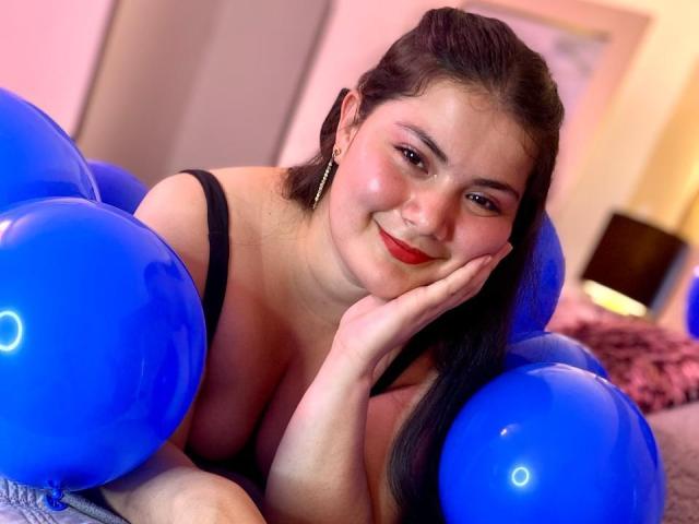 ZoeyClay, con globos azules, sonriendo, tiene lápiz labial rojo, cabello negro, y una mano debajo de la barbilla, vistiendo una blusa negra, mostrando sus senos, en un ambiente interior acogedor..