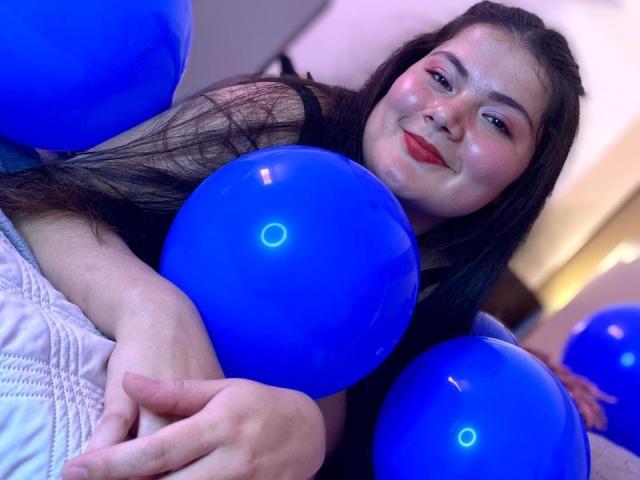 ZoeyClay com balões azuis, segurando um na mão direita, sorrindo para a câmera, cabelo longo e escuro, batom vermelho, cobertor branco acolchoado, balão azul na mão esquerda..