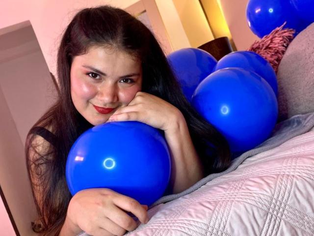 ZoeyClay, con cabello negro largo, sosteniendo globos azules, sonriendo con lápiz labial rojo, apoyando su barbilla en su mano, acostada en un sofá gris y texturizado..