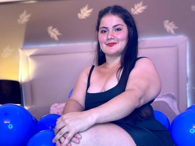 ZoeyClay, una mujer de talla grande con cabello negro largo, vestida con un vestido negro, sentada sobre globos azules, sonriendo, mostrando sus senos, con lápiz labial rojo y un rostro lindo..