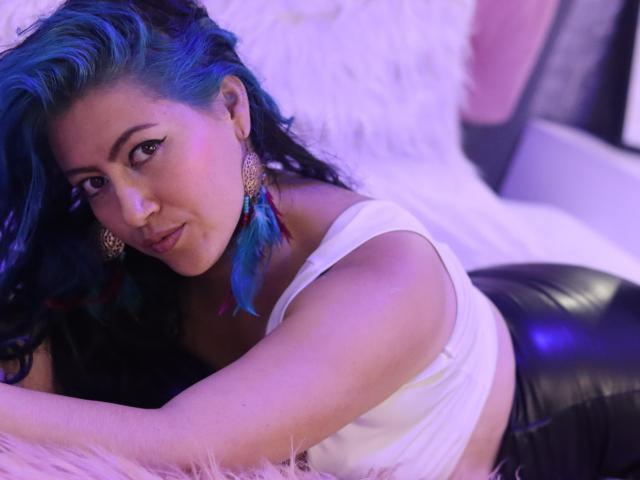 Ailich with blue and black hair, white tank top, and black leather pants, lying on a fluffy pink surface, looking at the camera with a slight smile.
