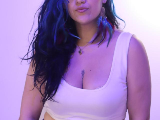 Ailich with blue-tinted wavy hair, white tank top, and necklace, showing her large tits and a small tattoo under her cleavage, against a purple-lit background.