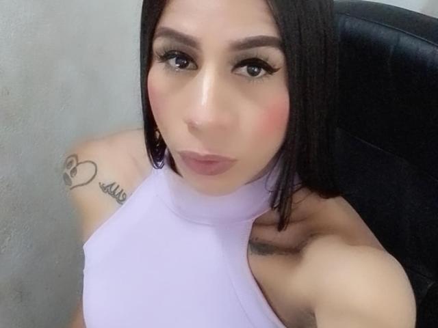 TrayXibi with a pouty face, wearing a lilac halter top, black hair, tattoo on right shoulder, large eyelashes, pink blush, and purple earrings, sitting in a black chair.