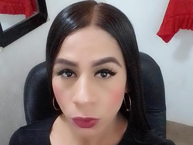 TrayXibi has dark straight hair, thick eyebrows, long black eyelashes, pink blush, red lipstick, and wears gold hoop earrings. She's sitting in a black chair, looking at the camera.