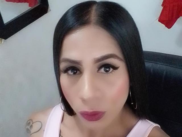 TrayXibi with straight black hair, pink lipstick, and hoop earrings, looking at the camera, wearing a pink top, showing a small tattoo on her right shoulder.