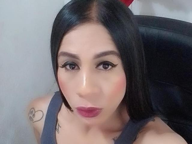TrayXibi has straight black hair, dark eyeliner, pink lipstick, and a tattoo on her left shoulder. She's wearing a gray tank top, looking at the camera.