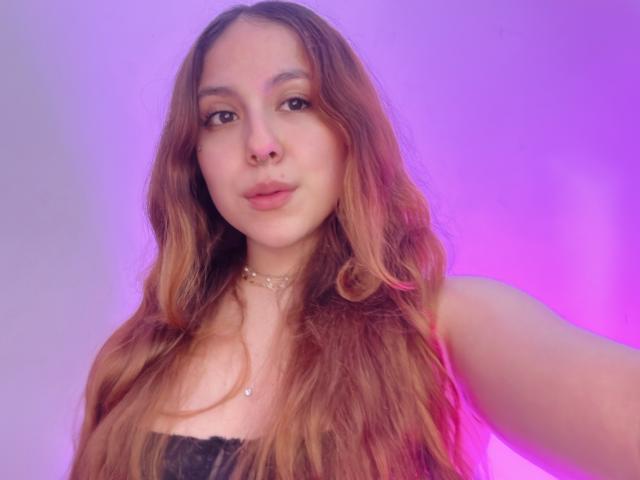 AmberlStha with long, wavy brown hair, wearing a black lace top, has a nose ring and a choker, with pink and purple lighting in the background.